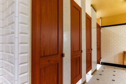 A row of wooden restroom stall doors with white tiled walls and floor in a well-lit bathroom.