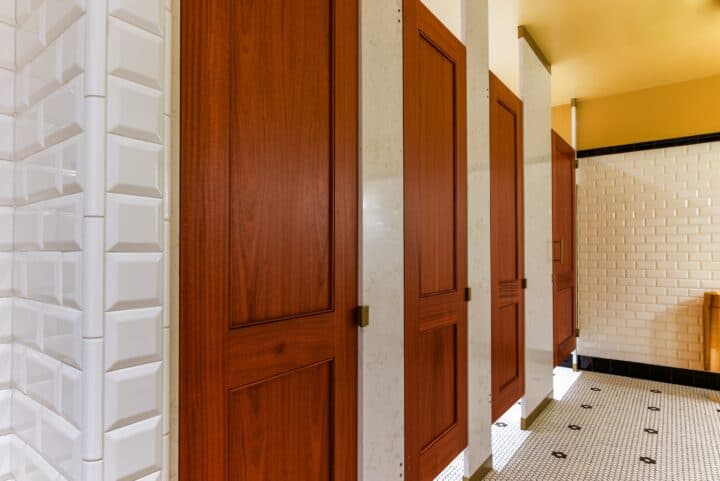A row of wooden restroom stall doors with white tiled walls and floor in a well-lit bathroom.