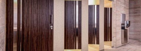 Modern public restroom with multiple closed stalls featuring dark wood-patterned doors, beige dividers, and a stainless steel urinal on the right side.