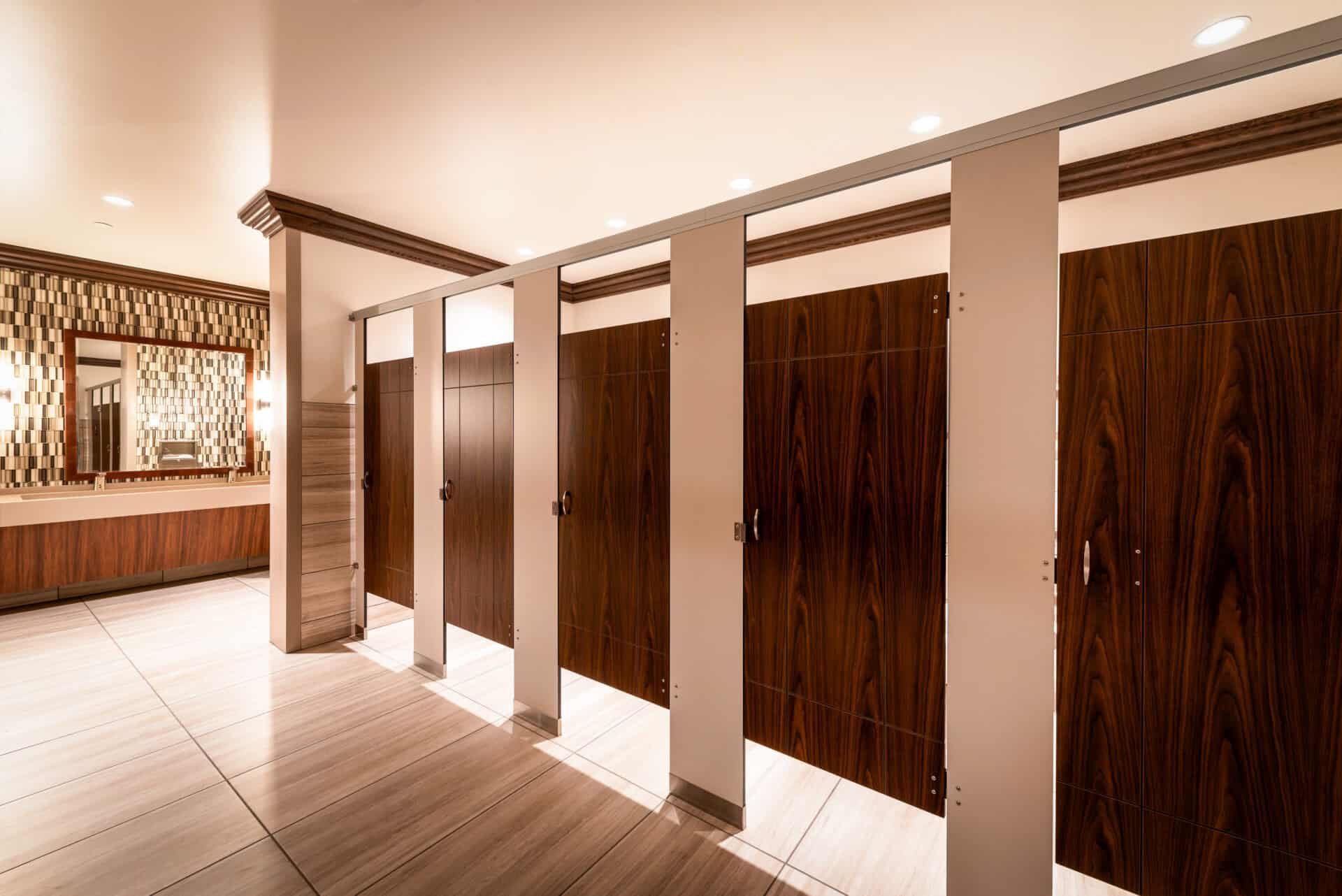 Commercial restroom stalls with engraved woodgrain compact laminate doors and contrasting light-colored pilasters in a headrail-braced configuration.