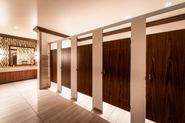 Commercial restroom stalls with engraved woodgrain compact laminate doors and contrasting light-colored pilasters in a headrail-braced configuration.