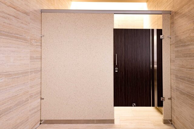 Modern public restroom stall with a wooden door and beige stone walls.