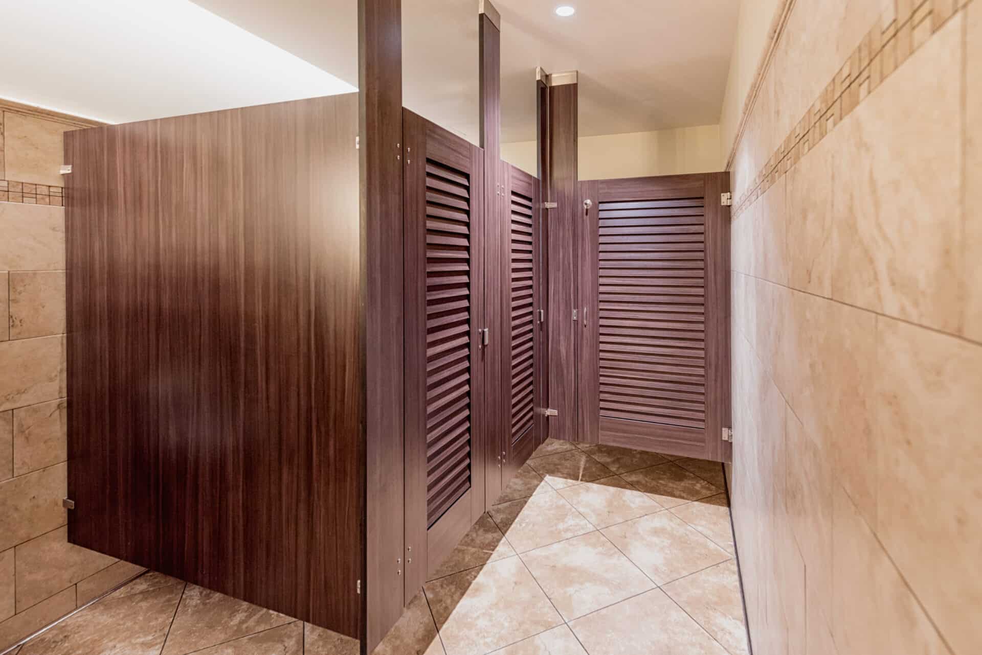 A clean restroom features stalls with wooden doors, a tiled floor, a granite countertop with a sink, and framed pictures on the wall.