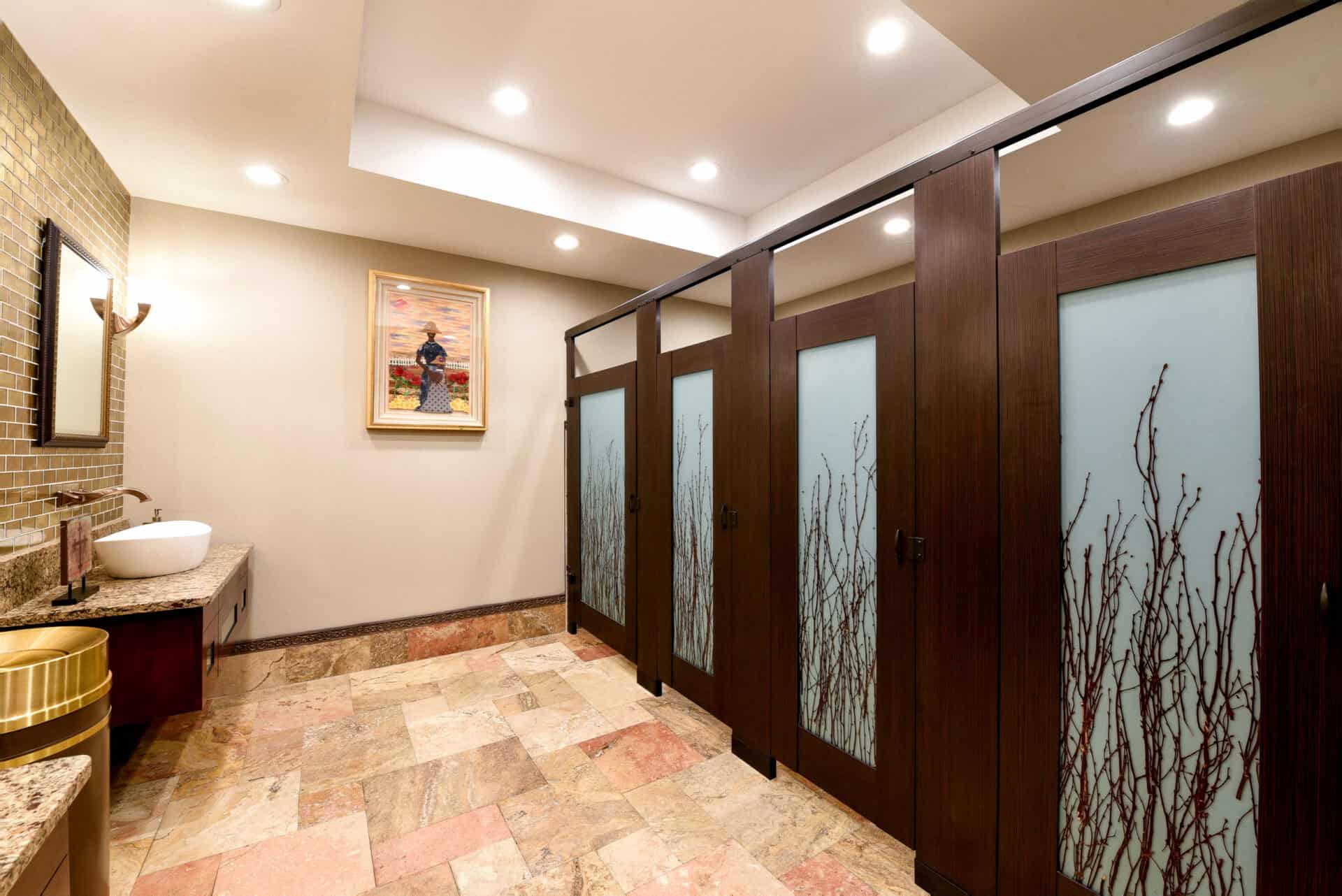 Restroom with woodgrain partitions featuring decorative frosted door lites with branch motif, paired with a custom vanity and warm tile flooring.