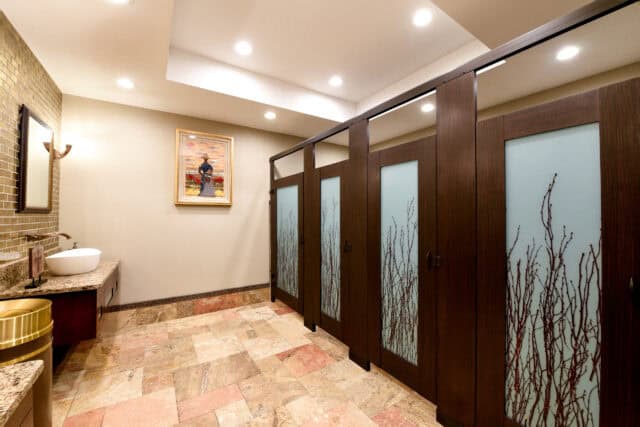 Restroom with woodgrain partitions featuring decorative frosted door lites with branch motif, paired with a custom vanity and warm tile flooring.