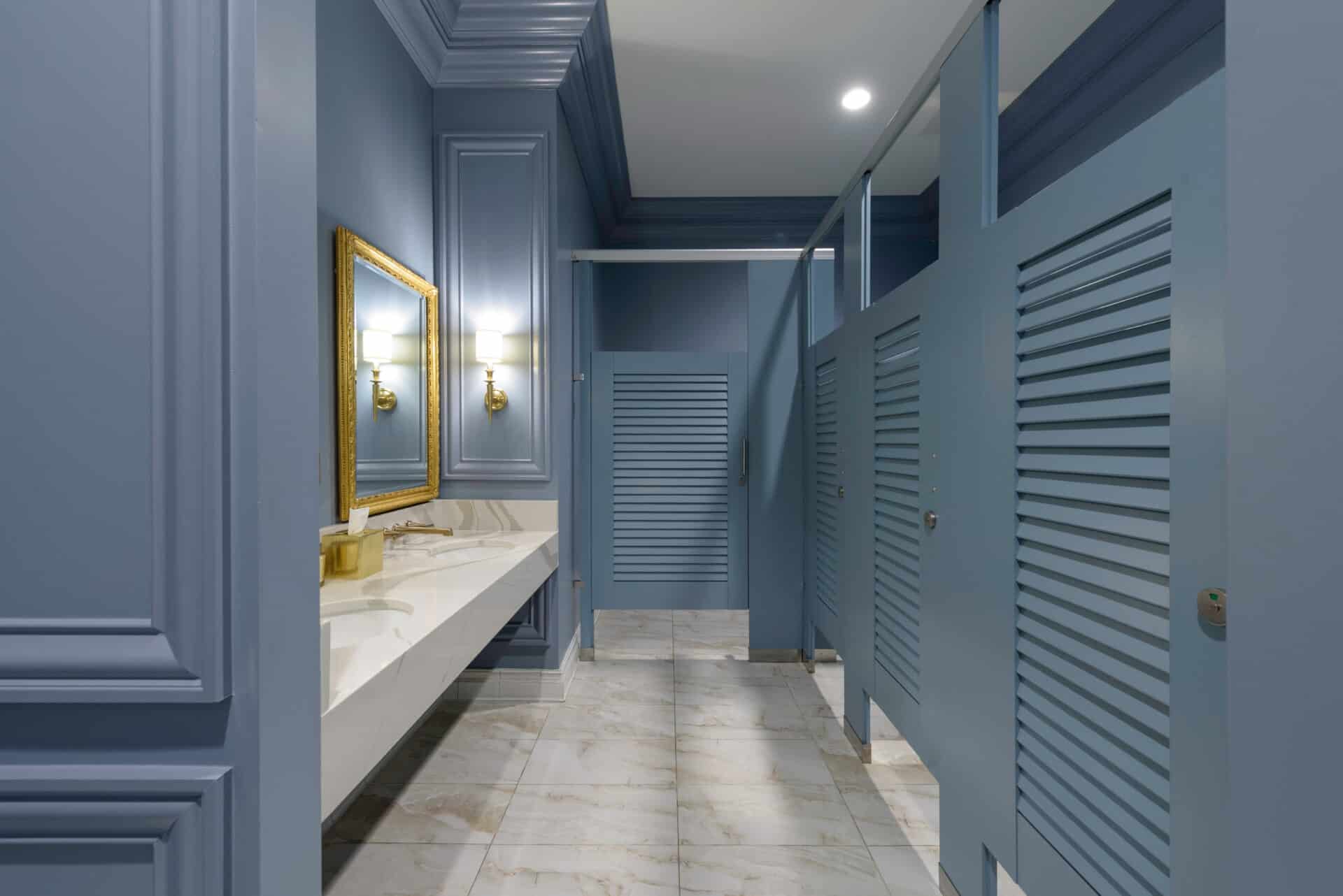 A public restroom with blue painted walls, a white marble floor, a counter with two sinks, a large mirror, and several stall doors on the right.
