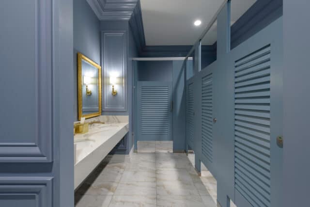 A public restroom with blue painted walls, a white marble floor, a counter with two sinks, a large mirror, and several stall doors on the right.