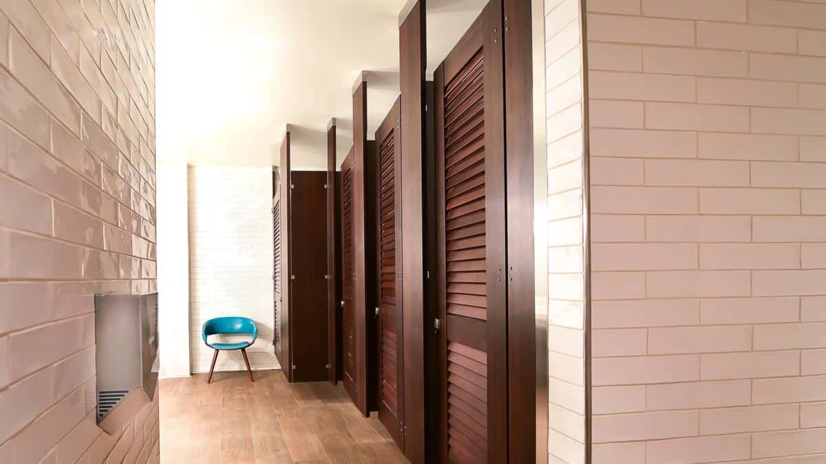 Row of dark wood louvered restroom partitions in a commercial bathroom with ceiling-hung mounting, white subway tile walls, and wood-look flooring.