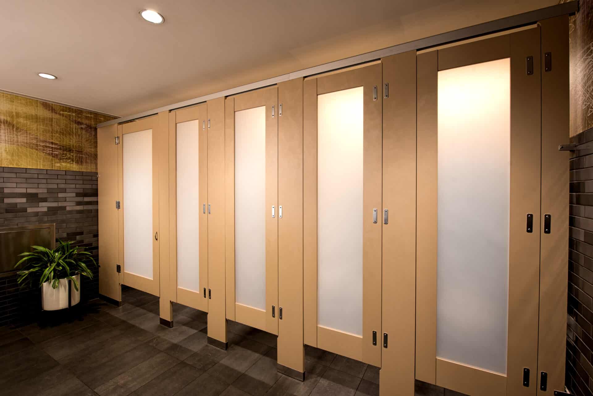 A row of beige commercial restroom partitions with frosted panels, dark tiled floor, and a potted plant in the corner offers a stylish privacy solution.