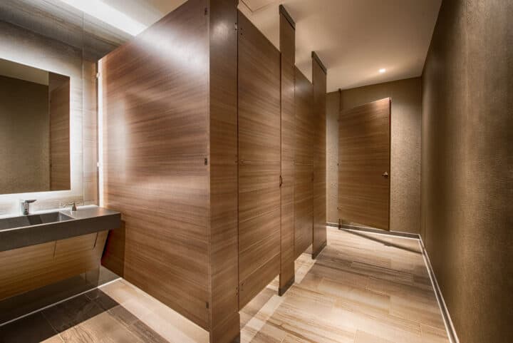 Public restroom with wooden partitions, a single sink with a mirror on the left, and brown tile flooring.