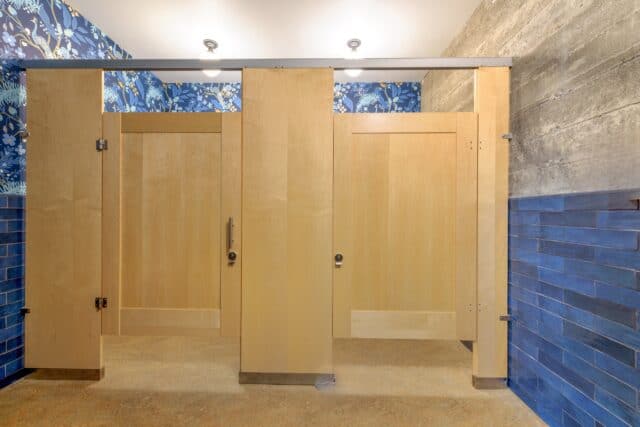 Public restroom with three wooden stalls, blue tiles, floral wallpaper, and overhead lighting.