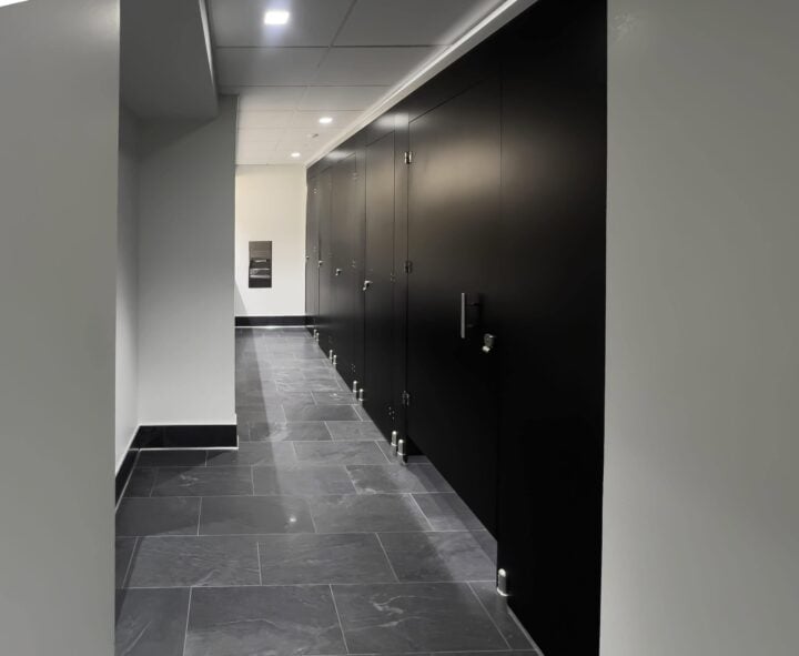 A hallway with a row of sleek, black European bathroom partitions on one side and a gray tiled floor. Ceiling lights illuminate the area.