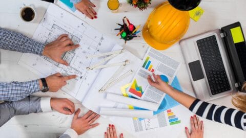 Team of professionals discussing commercial restroom designs with blueprints, charts, and a laptop on the table.