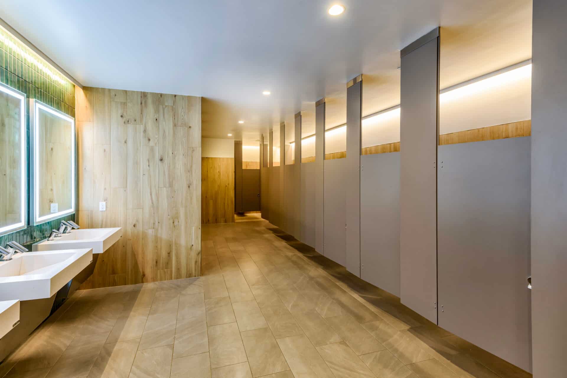 Contemporary commercial bathroom interior featuring gray bathroom stalls, wood-look wall paneling, floating white sinks, green tile accent wall, and LED-illuminated mirror