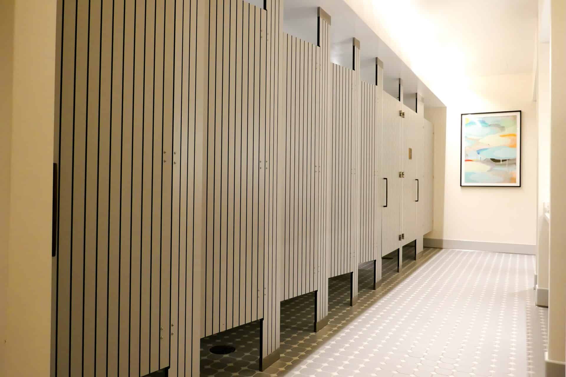 A row of compact laminate floor to ceiling mounted toilet partitions with engravings, a tiled floor, and a picture on the wall.