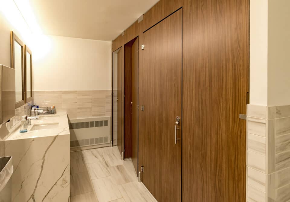 A public restroom with marble sinks and high privacy compact laminate woodgrain stalls, with a well-lit flat panel mirror above the counter.