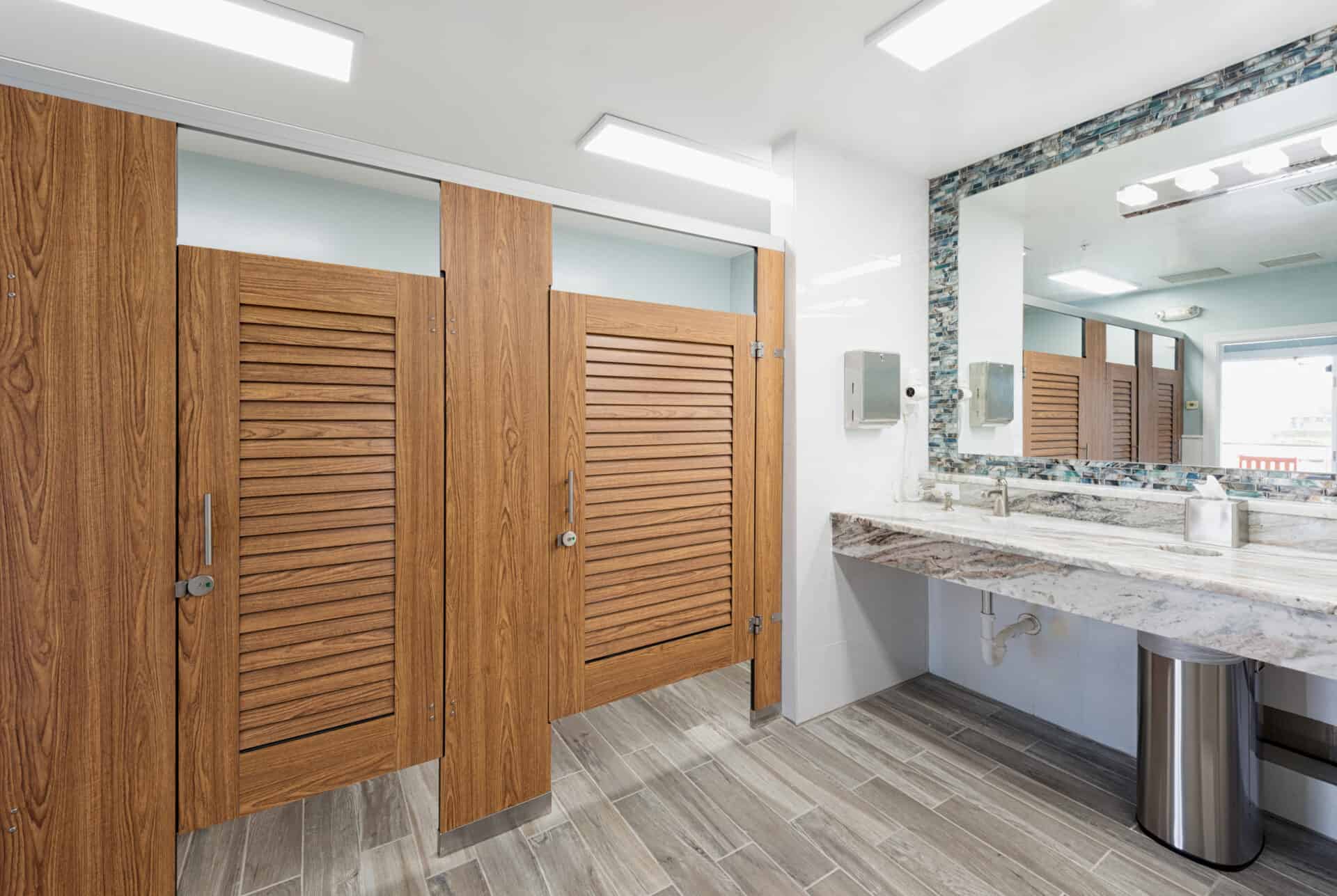 A public restroom with woodgrain compact laminate louvered stall doors, headrail braced mounting, a tiled floor, and a large mirror above a marble countertop with sinks.