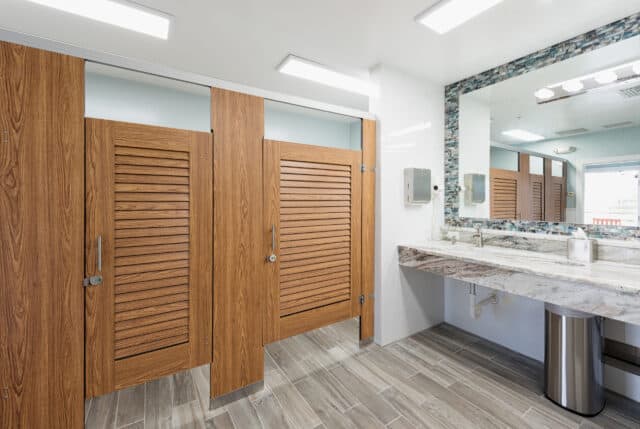 A public restroom with woodgrain compact laminate louvered stall doors, headrail braced mounting, a tiled floor, and a large mirror above a marble countertop with sinks.