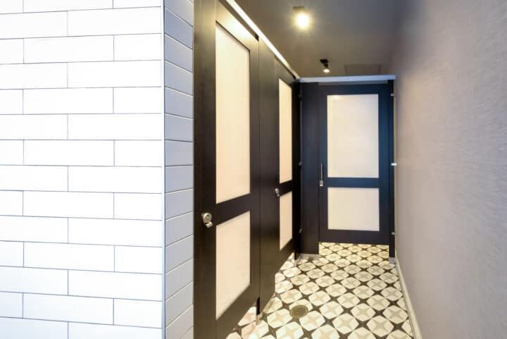 A compact laminate restroom partition with door lite panels, headrail braced for stability, and decorative geometric floor tiles.