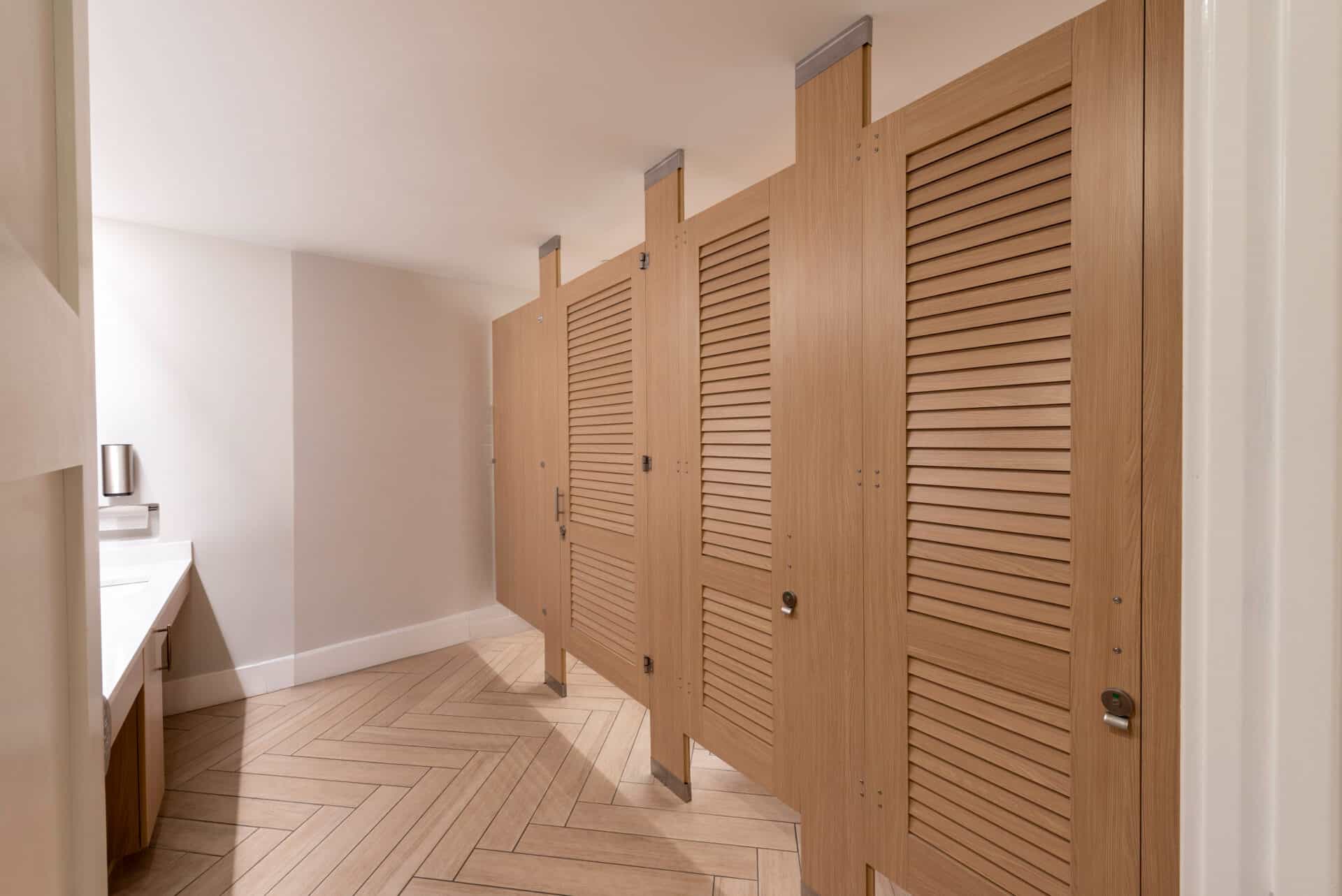 A clean public restroom with wooden stall doors, a tiled floor in a herringbone pattern, and a white countertop with a sink.