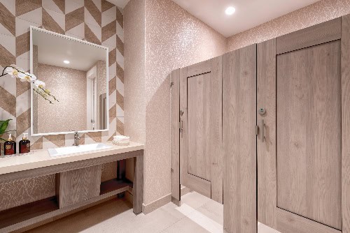 Modern restroom with a sink and large mirror on the left, and wooden door stalls on the right. Decor includes patterned walls and an orchid near the sink.