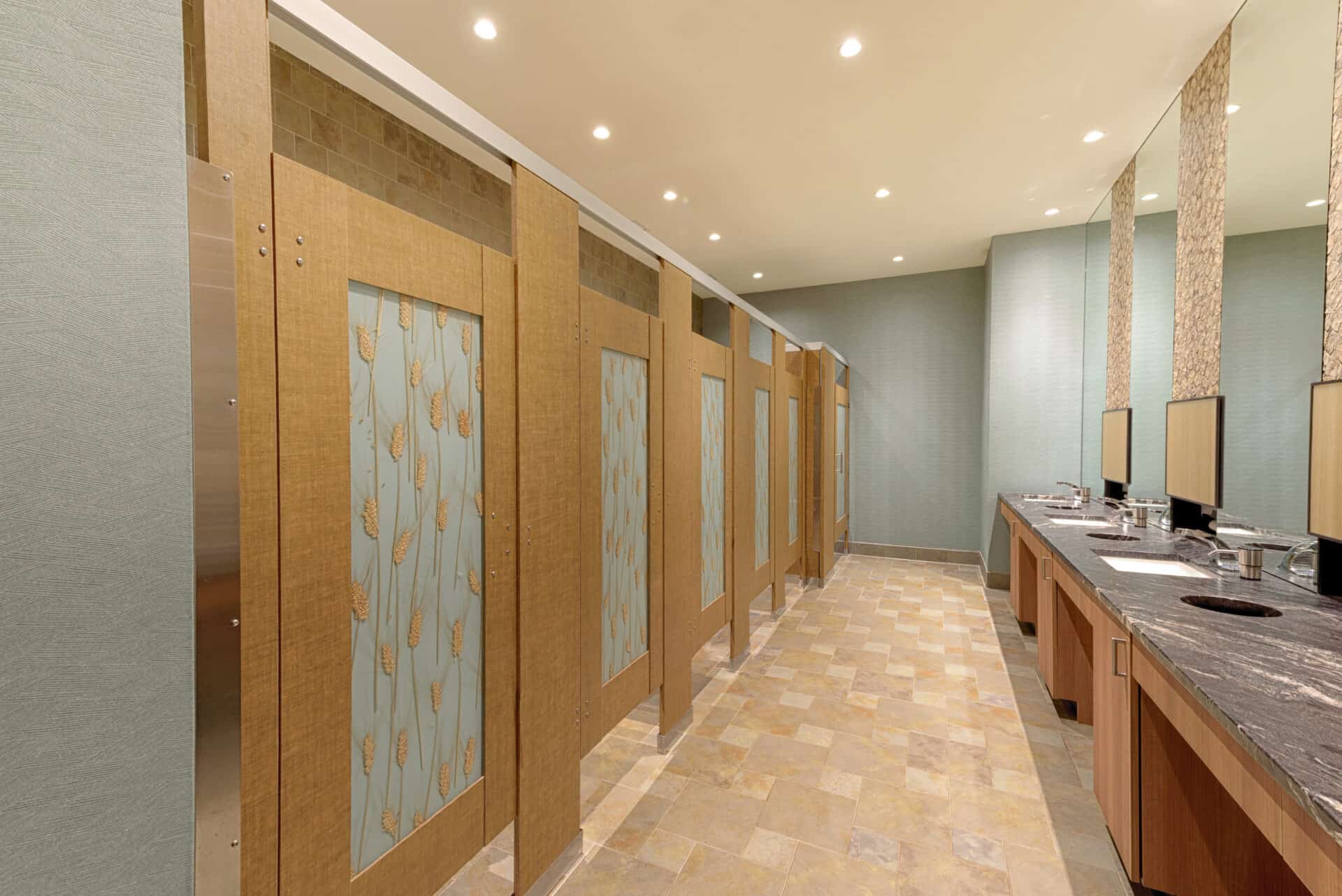 Restroom stalls with wood grain partitions featuring decorative glass panels embedded with wheat motif, set against tiled floors and a softly textured wall.