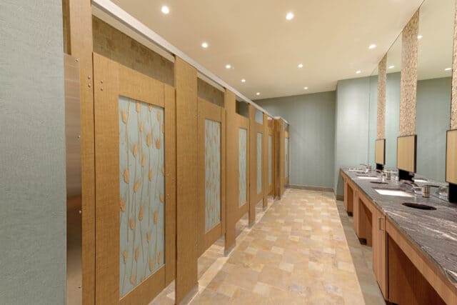 Restroom stalls with wood grain partitions featuring decorative glass panels embedded with wheat motif, set against tiled floors and a softly textured wall.