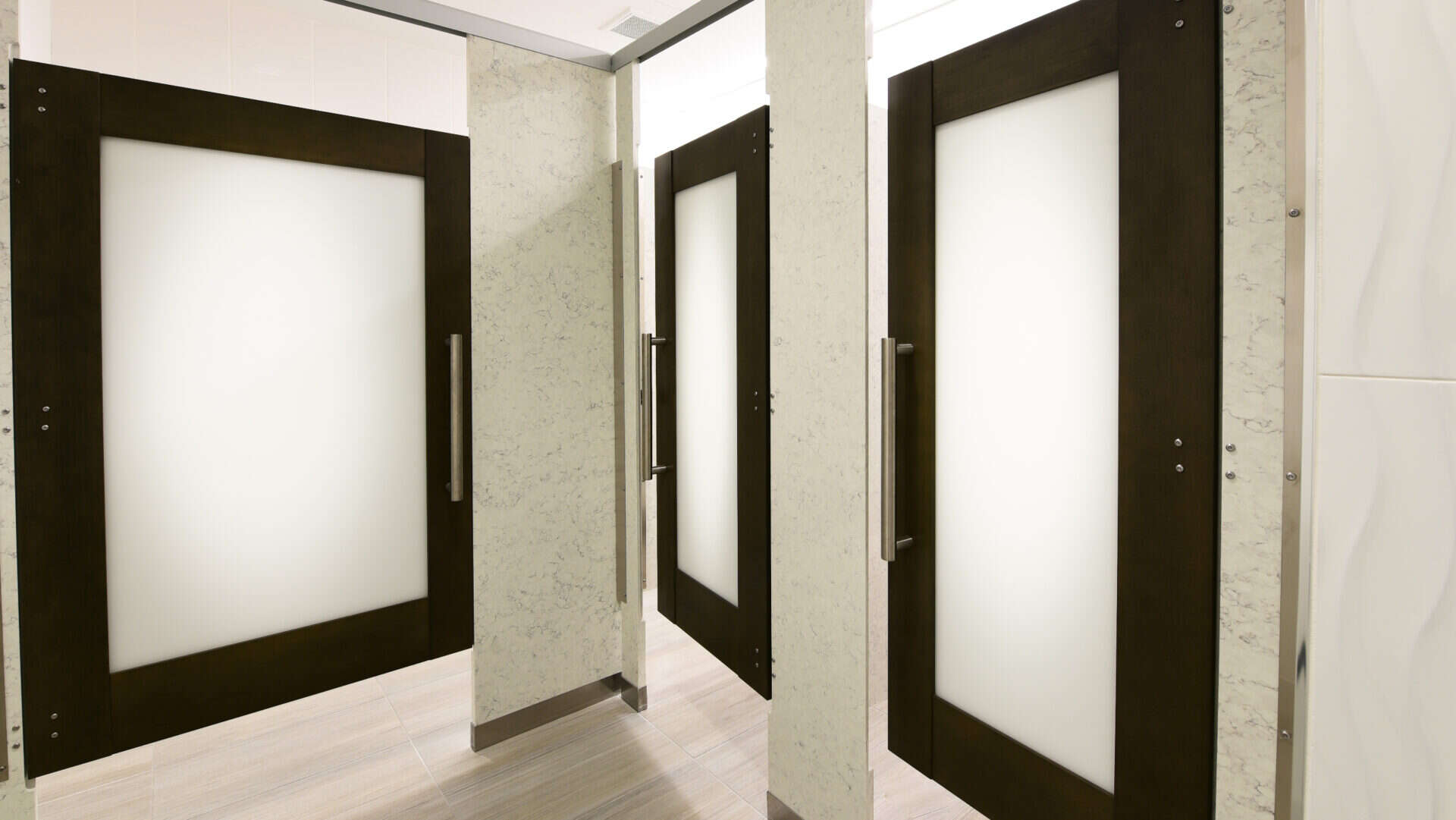 Three closed bathroom stall doors with frosted glass panels and metal handles in a tiled restroom.