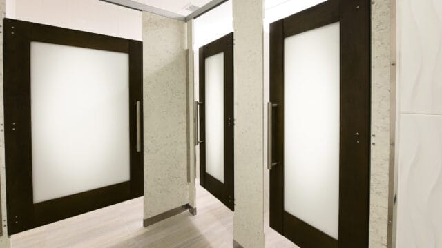 Three closed bathroom stall doors with frosted glass panels and metal handles in a tiled restroom.