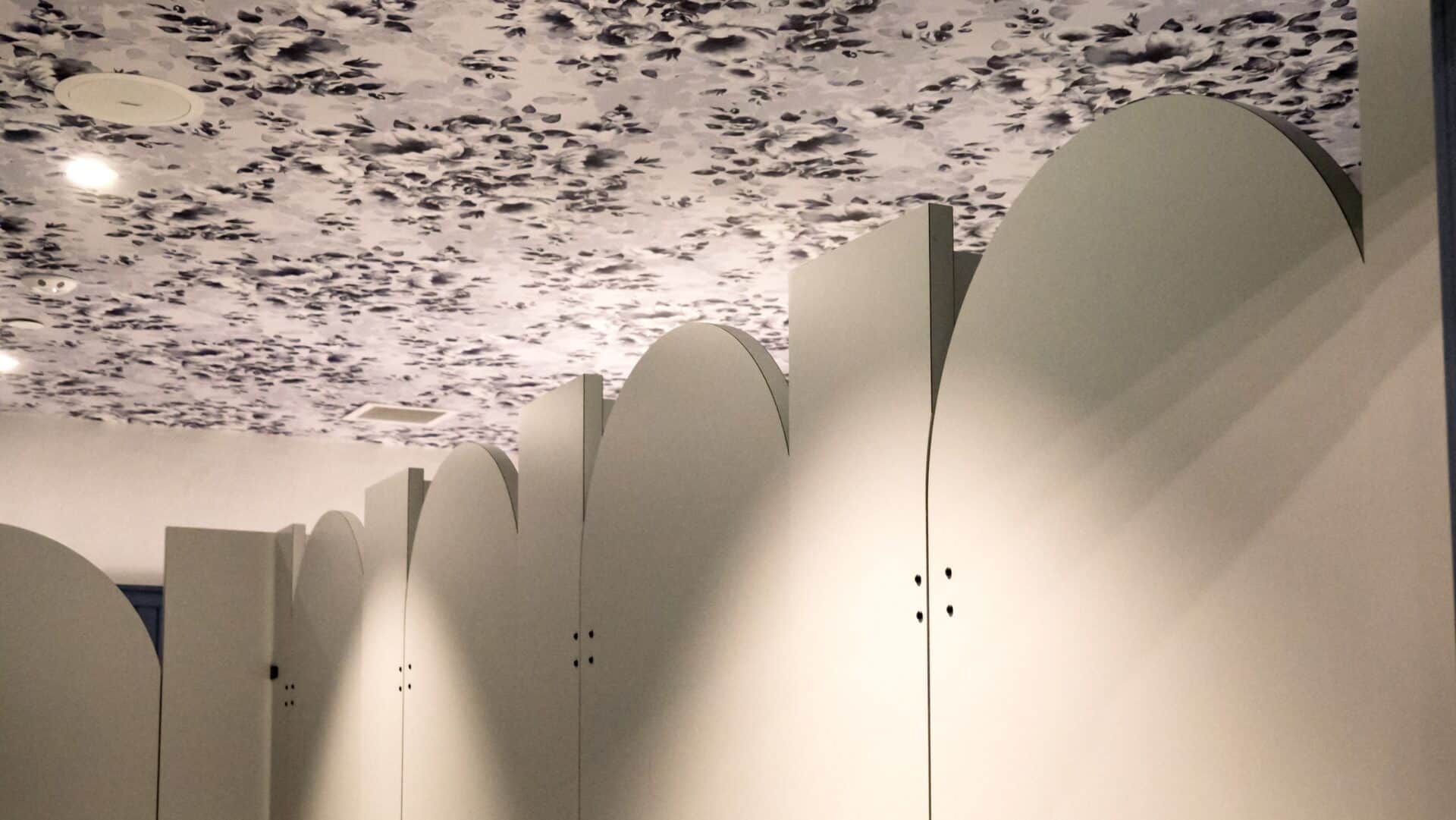Custom shape commercial restroom stalls with rounded door tops in a white plastic laminate. Bathroom stalls with wavy, white partitions and a floral-patterned ceiling.