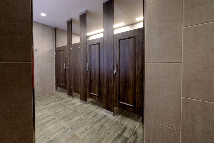 Restroom showcasing compact laminate partitions in dark wood finish with decorative molding and sleek tile flooring.
