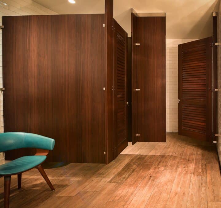 Ceiling hung restroom partitions with a dark wood veneer finish and louvered doors.
