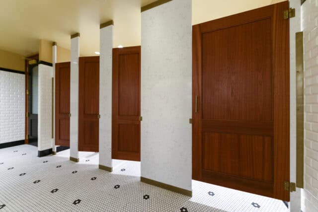 A public restroom with five wooden stall doors and white tiled walls and flooring.
