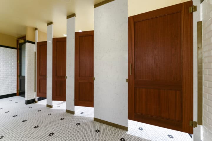 A public restroom with five wooden stall doors and white tiled walls and flooring.