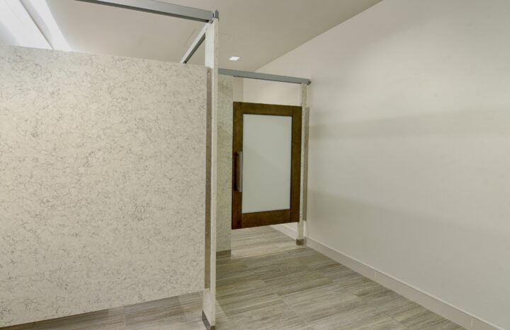 A restroom stall with engineered stone panels and wood veneer door lite doors with a frosted acrylic insert.