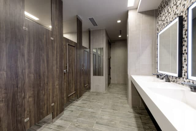 Modern restroom with illuminated mirrors, white sinks, and wooden stall doors. Walls and floor have neutral tiles.