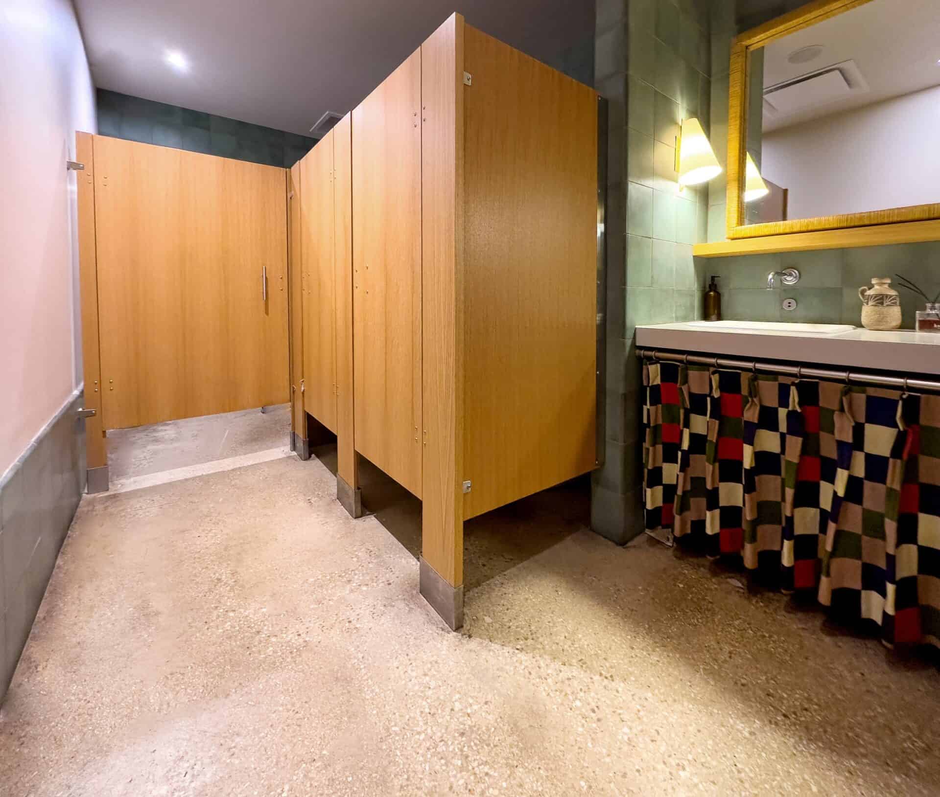 Commercial restroom partitions made from light wood veneer with stainless steel floor supports and floor mounting hardware.