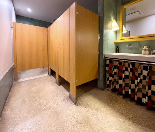 Commercial restroom partitions made from light wood veneer with stainless steel floor supports and floor mounting hardware.