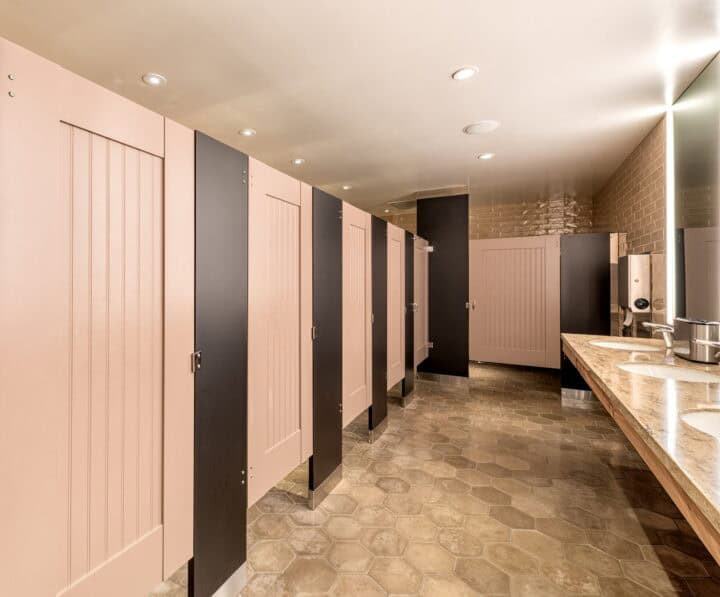 A clean public restroom with pink stall doors, beige tile flooring, and a long counter with sinks and mirrors.