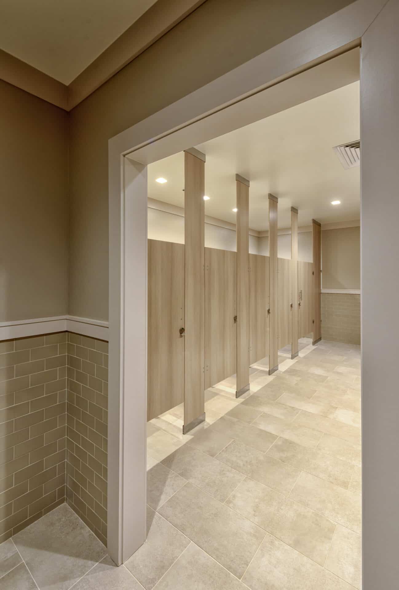 Public restroom interior with beige tiled walls and floors, featuring a row of light brown, closed stalls under bright overhead lighting.