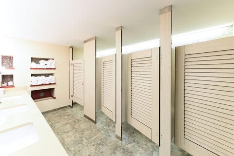 The public restroom features four light wood toilet partitions, a row of sinks with mirrors on the left, and shelves holding white towels.