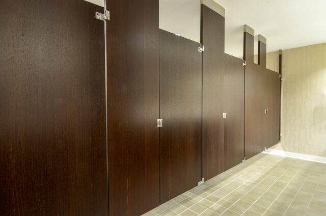 Row of closed wooden restroom stalls with beige tiled floor and ceiling lights.