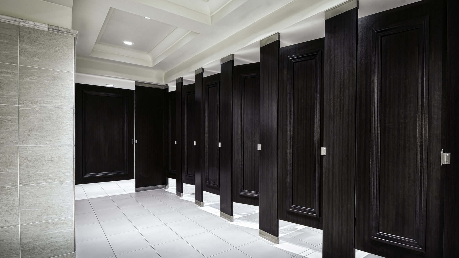A row of dark wooden restroom stalls with closed doors in a clean, modern public bathroom with white tiled floors and walls.