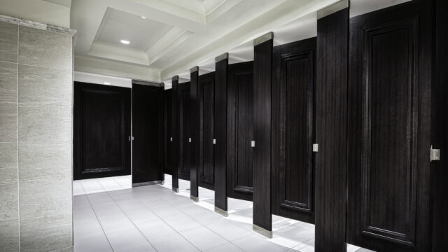 A row of dark wooden restroom stalls with closed doors in a clean, modern public bathroom with white tiled floors and walls.
