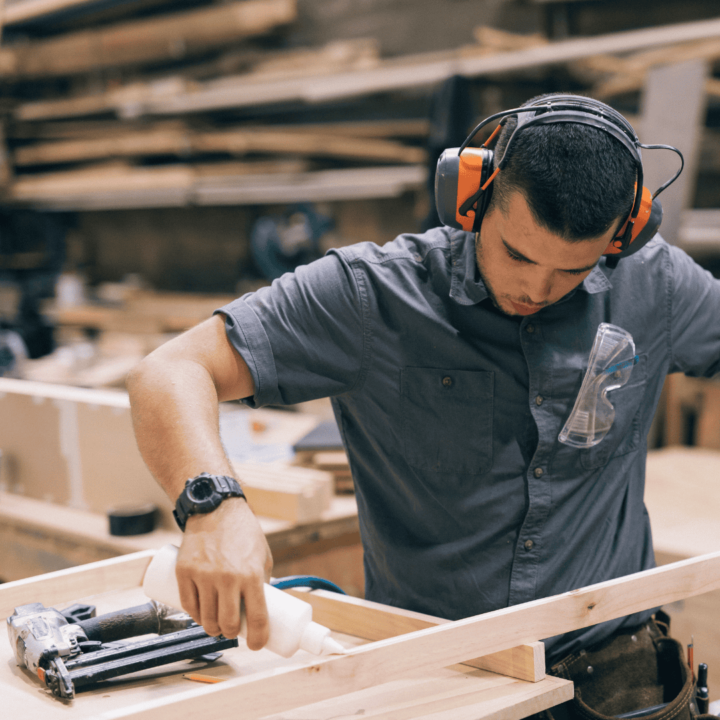 In a workshop, wearing ear protection and a gray shirt, a person meticulously applies adhesive to a wooden frame. Surrounded by various tools and boards, they demonstrate an understanding of different types of materials in their craft.
