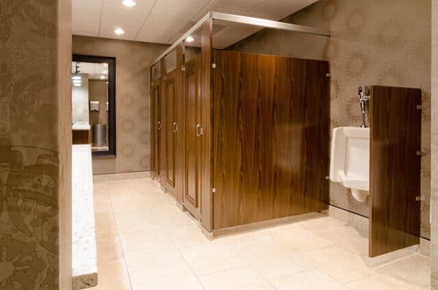 A clean public restroom with three wooden-door stalls, a white urinal with a privacy panel, and a sink with a mirror in the background. Beige tiled floor and patterned walls are also visible.