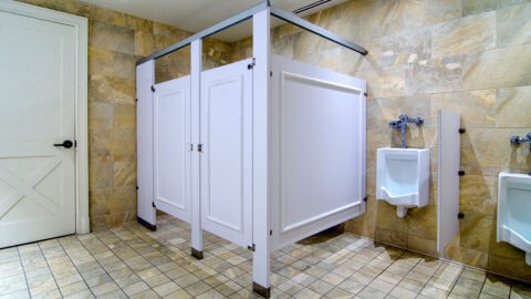 Public restroom featuring two Ironwood Element Series white stalls and two wall-mounted urinals on beige tiled walls and floor. A white door with a black handle is on the left, showcasing the high-quality craftsmanship of customizable restroom partitions.