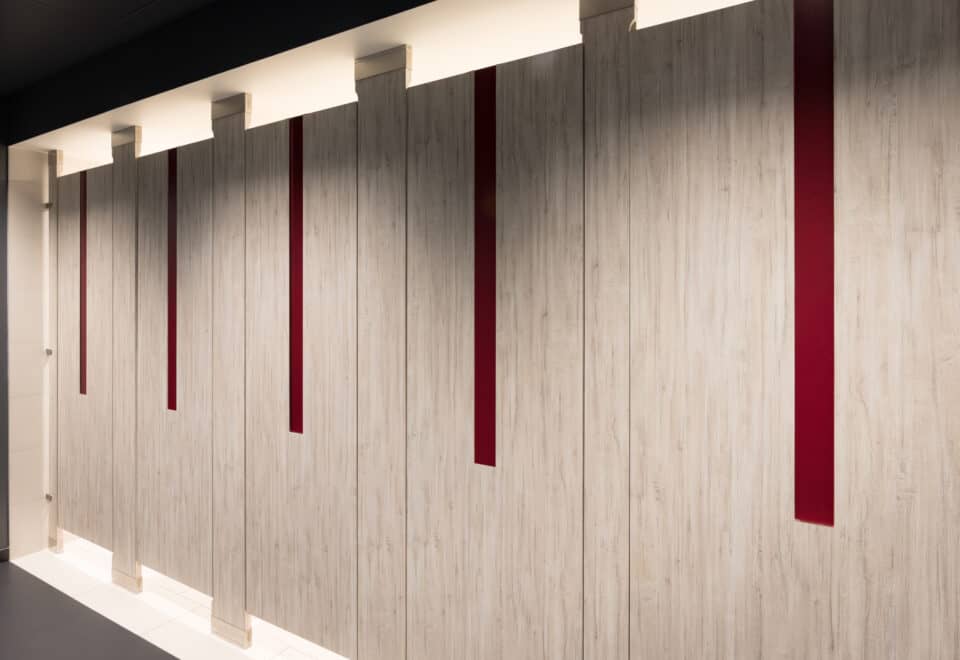 A commercial upscale restroom featuring partitions with high pressure laminate woodgrain doors and pilasters. Floor to ceiling mounted with red acrylic inlays in the doors.