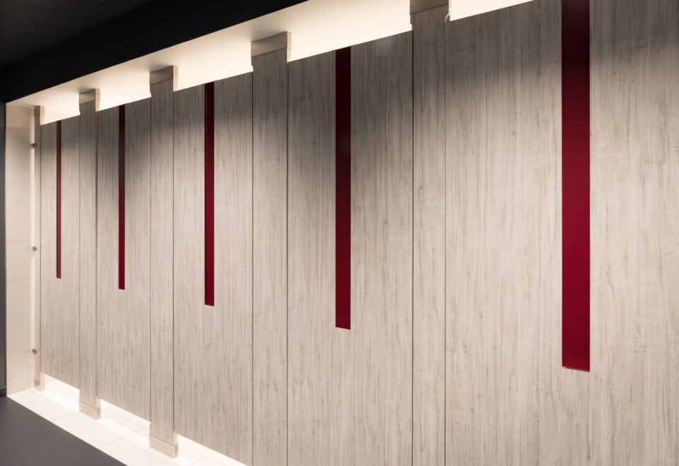 A commercial upscale restroom featuring partitions with high pressure laminate woodgrain doors and pilasters. Floor to ceiling mounted with red acrylic inlays in the doors.