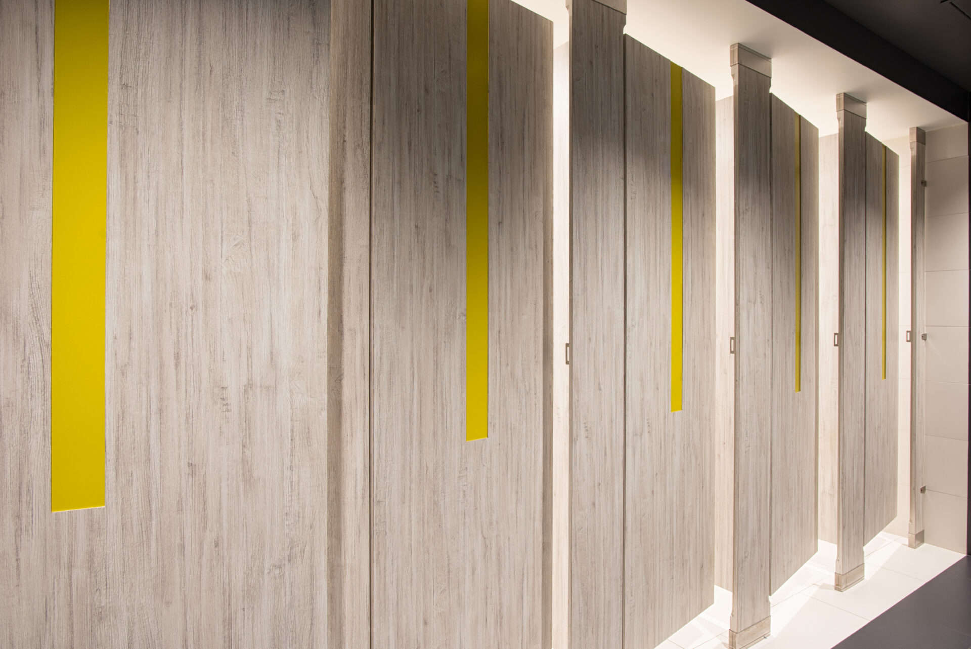 A commercial upscale restroom featuring partitions with high pressure laminate woodgrain doors and pilasters. Floor to ceiling mounted with yellow acrylic inlays in the doors.